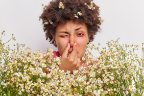 Headshot of curly haired young ethnic woman has problems with breathing holds nose suffers from allergy to camomile holds big bouquet of flowers has red itchy eyes. Allergic reaction on pollen